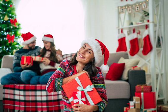 Merry Christmas and Happy New Year. Photo of a happy smiling beautiful woman in a Christmas Santa hat is showing a gift box in hands on group of friends and Christmas tree background.