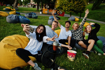 Young multi ethnic group of people watching movie at poof in open air cinema and making selfie on...