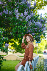 Naklejka premium Closeup young girl standing on a background of lilac bushes. she is of European appearance in a brown hat and dress