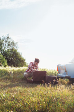 Retro-Styled Young Man Looking at Clothes in Suitcase on Vintage Car Summer Road Trip