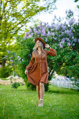 Obraz na płótnie Canvas Attractive stylish smiling slim girl with long hair walks in the park, dressed in a warm brown dress, autumn fashionable street fashion, with a hat. Young woman of European appearance