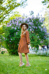 stylish girl in a brown hat and a light dress on a background of lilac lush bushes. Young woman with a smile on her face on a sunny summer day walks in the park