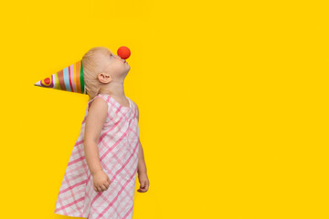 Little girl in party hat and red clown nose. Portrait side view on yellow background. Copy space