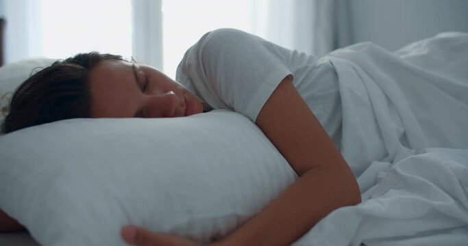 A young brunette woman wakes up in the morning in bed against the background of a large window. Portrait of a smiling woman. High quality 4k footage