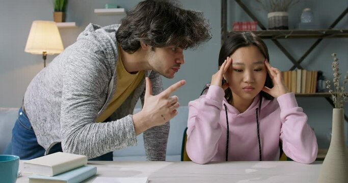 Angry caucasian guy shouting at his girlfriend during self isolation. East asian girl trying to calm down at table. Domestic violence, abusive relationship 4k video