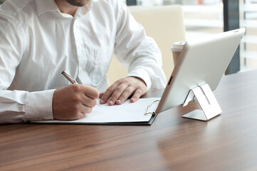 Businessman in a white shirt with a digital tablet in his hands signs a contract in the office. Workplace with a cup of coffee and a document with a pen on a wooden table.