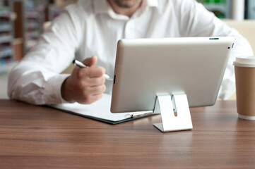 Businessman in a white shirt with a digital tablet in his hands signs a contract in the office. Workplace with a cup of coffee and a document with a pen on a wooden table.