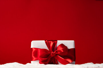 Christmas surprise. Holiday present. Wrapped white gift box with ribbon bow on snow pile isolated on red copy space festive background. New Year greeting card.