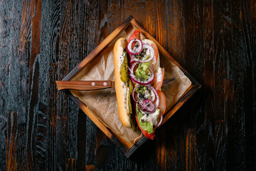 Hot dog with onion tomatoes cucumber and sauce on a wooden stand served with a knife. Fast food with restaurant serving.