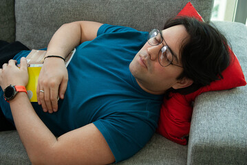 Close up portrait of a young man with glasses sleeping on a grey couch in the living room next to the window, holding a book between his hands.