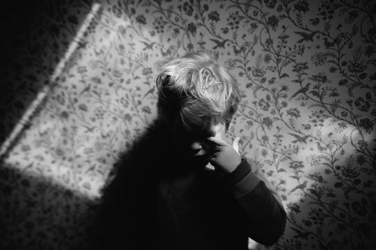 Young child rubbing her eye while standing in a patch of light on a wallpapered wall.