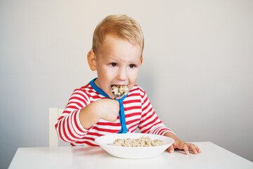 little boy 2 years old sits at the table and eats oatmeal with a large spoon. correct nutrition...