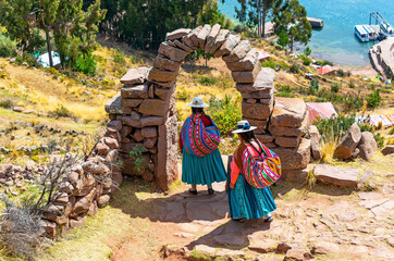 Two peruvian indigenous Quechua women in traditional clothing under an arch walking down steps to a...