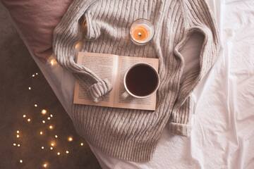 Cup of coffee or black tea staying on open book with burning candle and knitted gray sweater in bed...