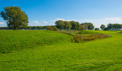 Dike in a green grassy field in sunlight under a blue sky in autumn, Almere, Flevoland, The Netherlands, September 24, 2020
