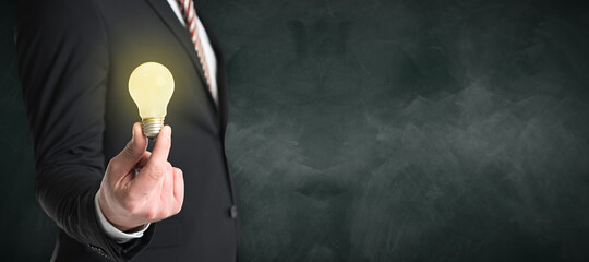 businessman holding a lit lightbulb in front of a chalkboard