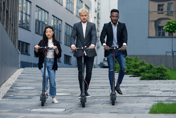 Fototapeta premium Attractive joyful multiracial office colleagues riding on electric scooters near beautiful architectural building.