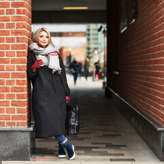 Outdoors lifestyle fashion portrait of stunning blonde girl. Drinking coffee and walking on the city street. Businesswoman.Wearing stylish black oversized coat, wide warm scarf and burgundy gloves.  