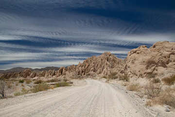 Fototapeta premium Road trip. Driving along the dirt road across the arid desert. The route, rocky valley and beige sandstone hills under a beautiful blue sky.