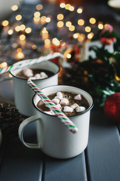 Two enamel mugs with hot chocolate and marshmallow on a table with festive lights in background.