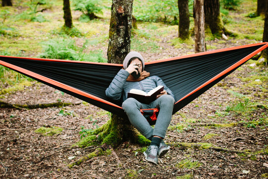 Young Man Sitting In A Hammock Reading A Book And Sipping Coffee From A Canteen Cup In the Woods