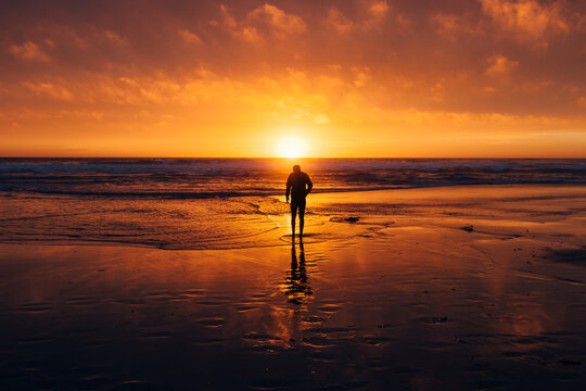 A Young Man Stands On The Shore As The Sun Sets Over The Pacific Ocean