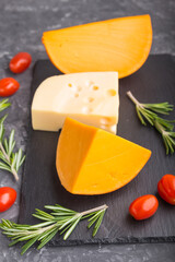 Various types of cheese with rosemary and tomatoes on black slate board on a black concrete background. Side view, close up.