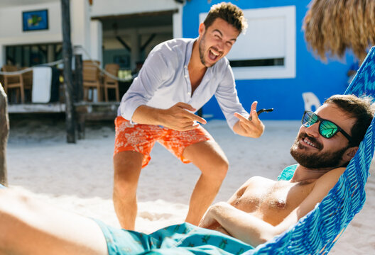Young man photobombing and bothering his friend who is resting on a hammock in the beach