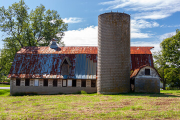Fototapeta na wymiar An abandoned old farm house with a tall brick silo and a big barn with rusted tin ceiling. This very old run down builsing complex is surrounded by grassland and trees. Image taken in rural Maryland.