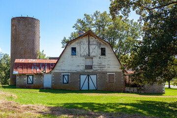 An abandoned old farm house with a tall brick silo and a big barn with rusted tin ceiling. This very old run down builsing complex is surrounded by grassland and trees. Image taken in rural Maryland.