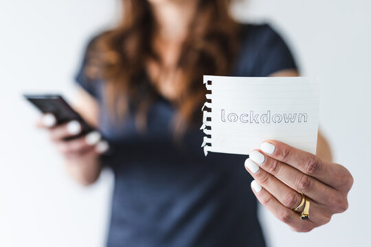 covid-19 pandemic outbreak, woman holding Lockdown text towards the camera while browsing on her smartphone shot at shallow depth of field - Powered by Adobe
