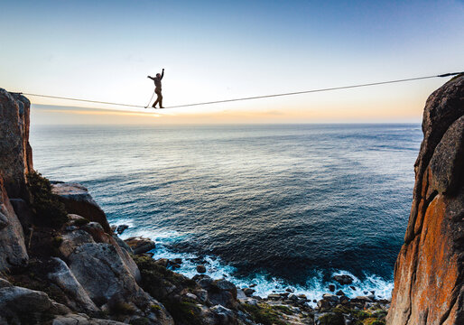 Man tightrope walking a highline between two cliffs overlooking the sea at sunset