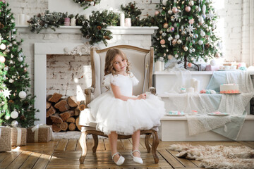little beautiful girl in a white fluffy, elegant dress is sitting in a chair on the background of Christmas trees in the interior of the loft. new year holidays and vacations.