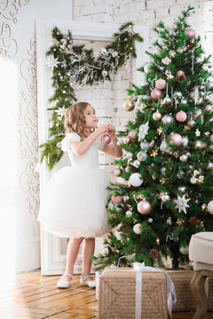 A Beautiful Little Girl In A White Dress Is Decorating A Christmas Tree. Home Decoration For New Year Celebration. Winter Vacation. Happy Childhood.