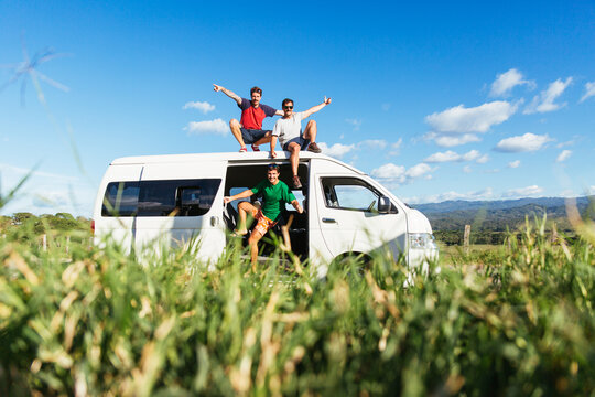 Three excited young men sitting on the roof of a white minivan during a break stopover in the middle of the road in the countryside with grass in front
