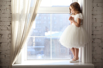 a beautiful little girl in a white dress is waiting for Santa Claus at the window. children's fairy...