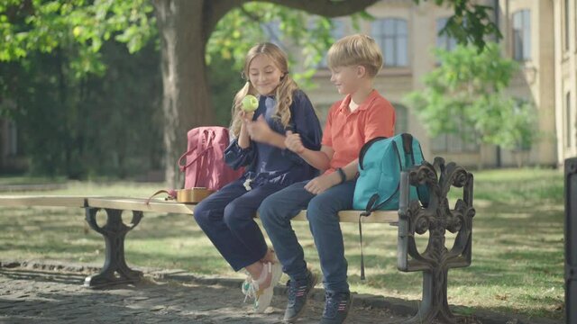 Wide shot of charming boy and girl sitting on bench at schoolyard. Cute Caucasian schoolboy giving healthful green apple to schoolgirl. Concept of first love and children flirt.
