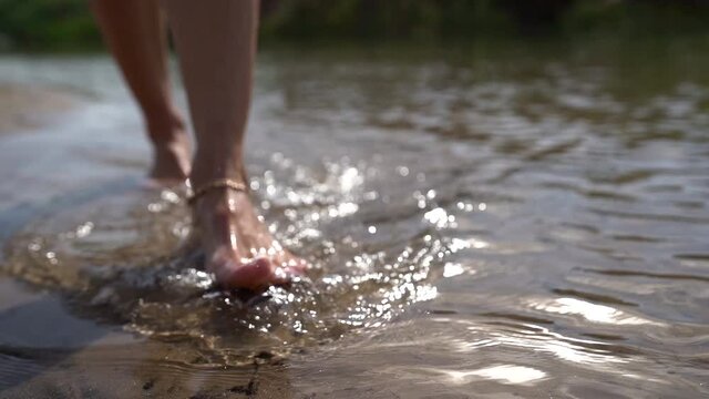 Close-up of women's bare feet with an ankle bracelet walking on the sandy shore of a wild forest lake in summer.