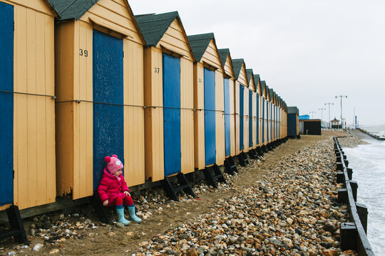 Little girl wearing a pink jacket sits outside a row of beach huts in winter.