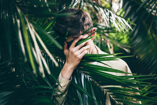 Young man hiding his face with hand while standing in palm trees forest