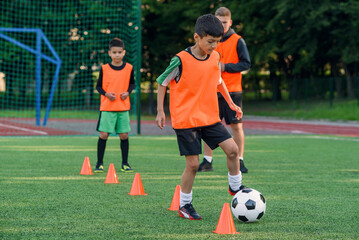 Obraz premium Persistent teen boy in sportswear trains football on soccer field and learns to circle the ball between training cones.