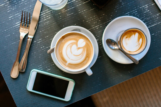 A cell phone laying on a breakfast table by a latte and espresso
