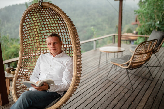 Tourist man swing on wicker rattan hang chair in the jungle, nature mountains view, hold in hands book,reading