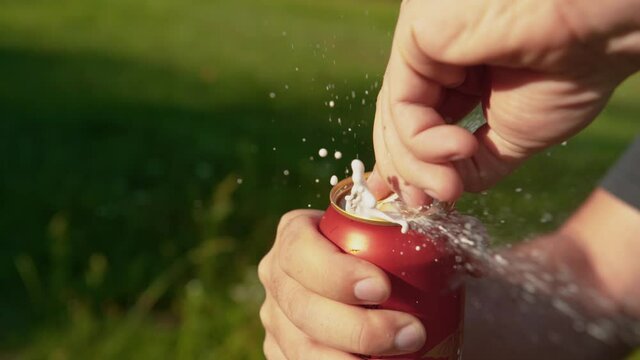 SLOW MOTION, CLOSE UP, DOF: Unrecognizable thirsty man opens a can of shaken up beer during picnic. Unfortunate guy spills refreshing cold beer while pulling the tab of a large red aluminum can.