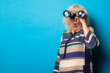 Surprised old woman in a striped dress looks through binoculars on a blue background