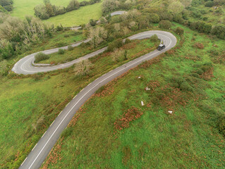 Small narrow country road in Burren, Ireland, Black bus moving down the hill, aerial drone view. S shape of the road.