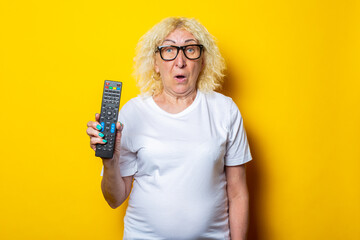 Surprised blonde old woman with glasses holding remote control on yellow background.