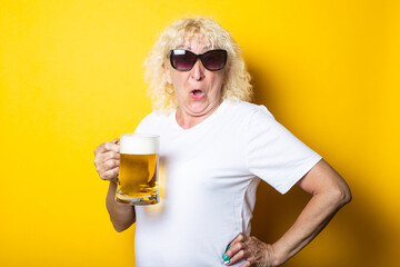 Surprised blonde old woman in a white T-shirt, in sunglasses with a glass of beer on a yellow...