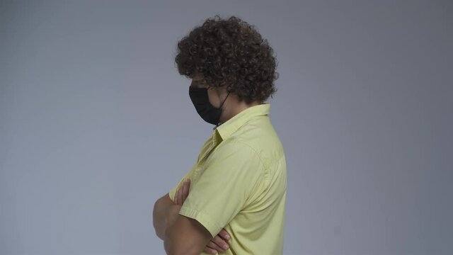 Curly man stands in a profile in a medical mask. Guy crossed his arms on his chest and turns discontentedly into the camera. High quality.