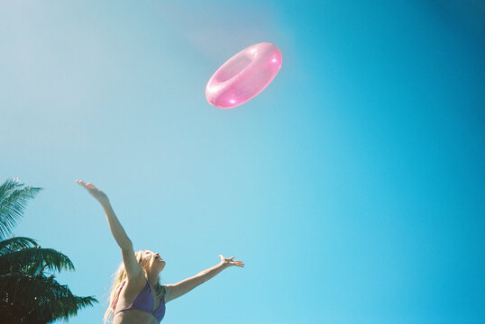 blonde girl in bikini throwing pink floatie in air against blue sky